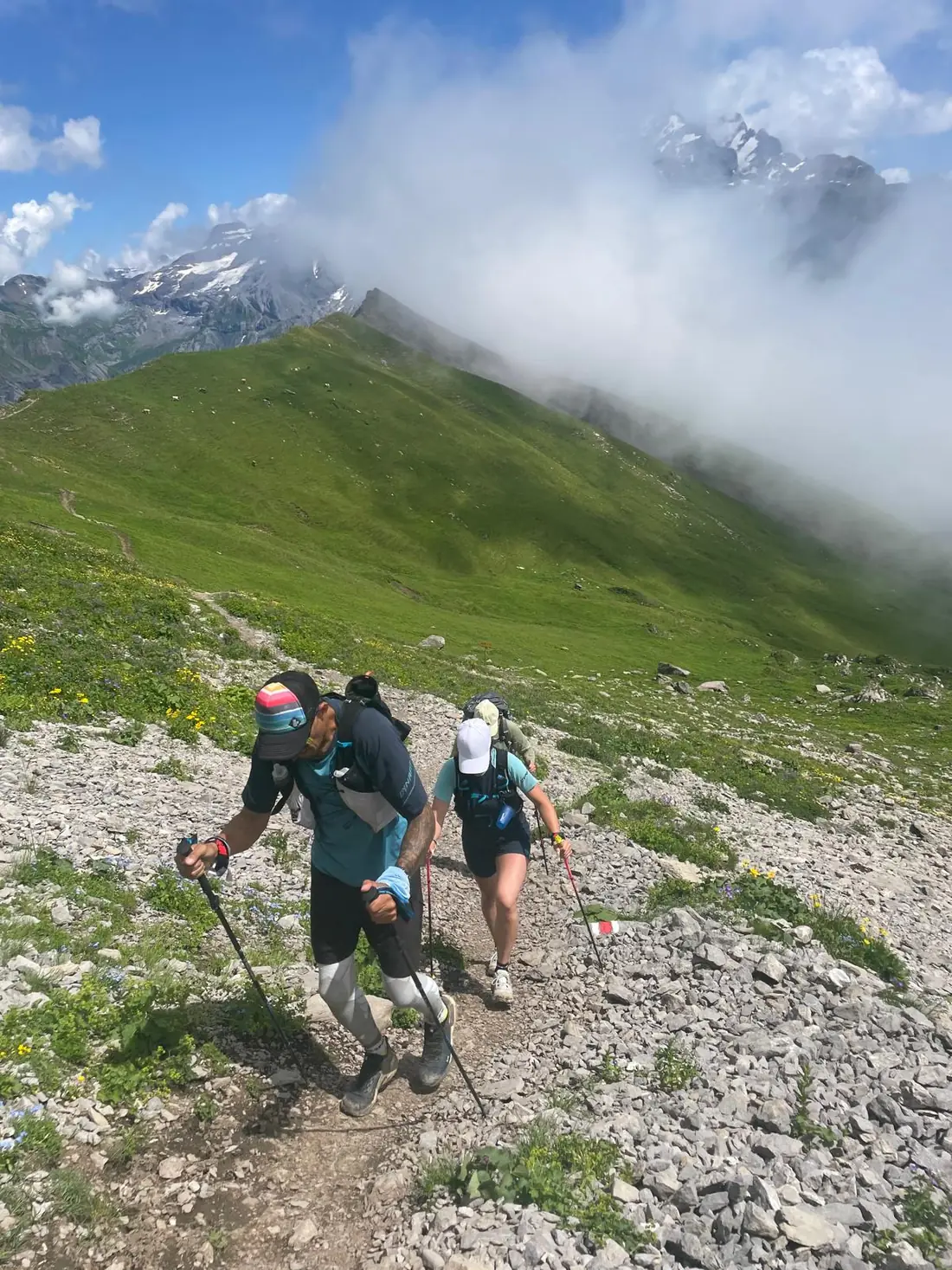 Deux traileurs, bâtons en main, gravissent un sentier de crête dans les Alpes suisses, sous les nuages.