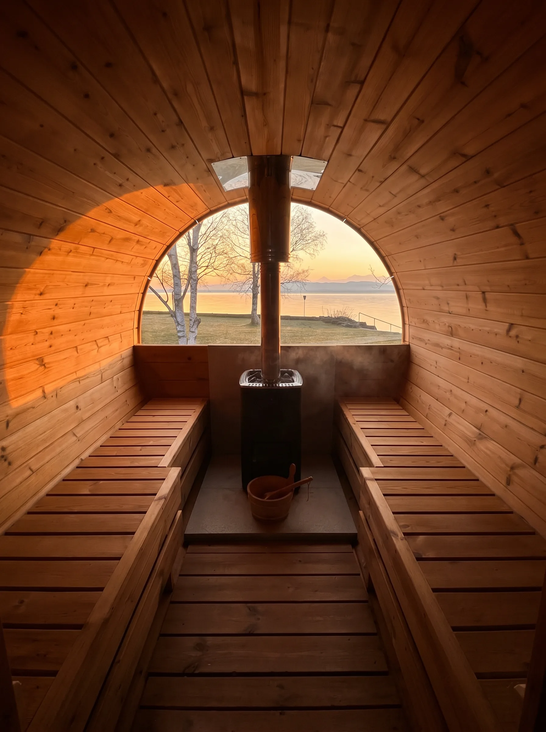 Intérieur d'un sauna tonneau en cèdre, poêle à bois et fenêtre arrondie ouverte sur le lac au coucher du soleil