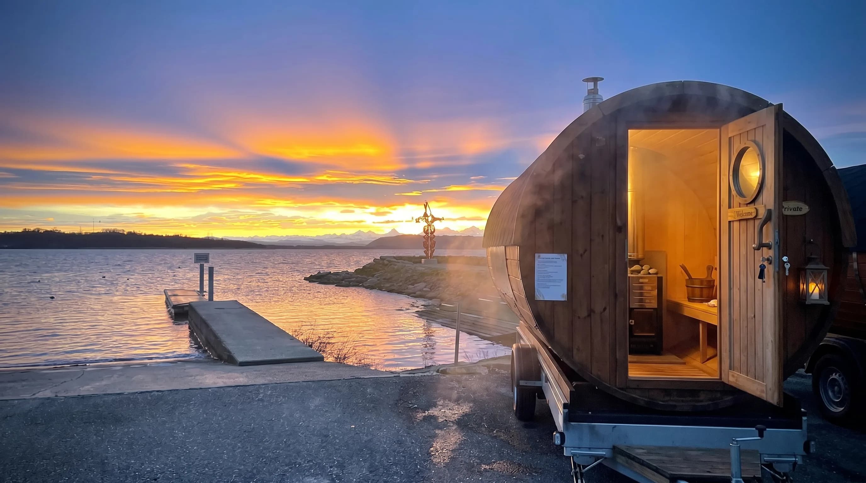 Sauna tonneau au bord du lac à la tombée du jour, lumière chaude par la porte ouverte